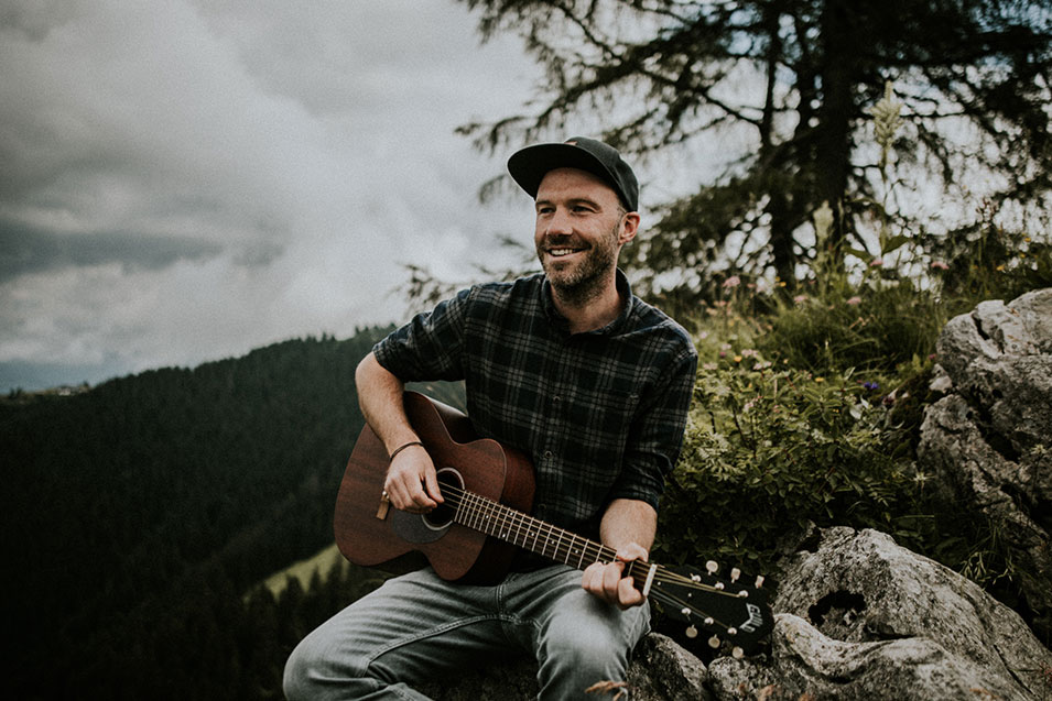 Komponist Michael Regner spielt Gitarre im Freien und sitzt auf einem Stein am Waldrand